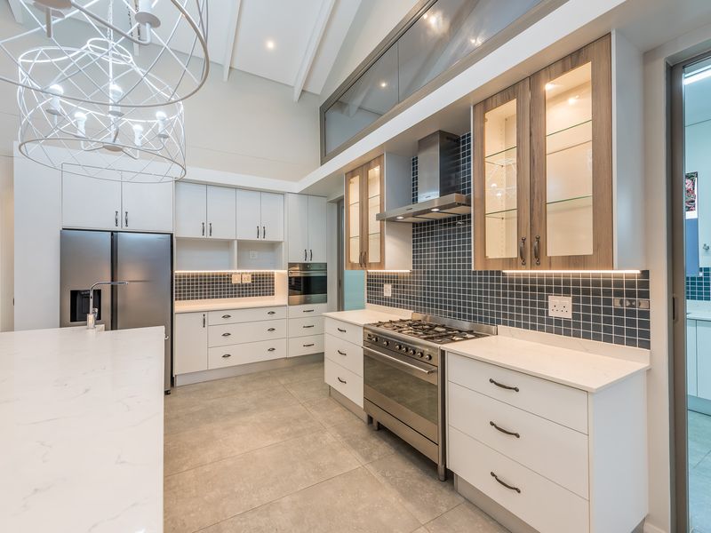 Modern kitchen featuring stone countertops, tiled backsplash, stainless steel gas range, extractor fan, and integrated cabinetry with under-cabinet lighting and high vaulted ceilings.