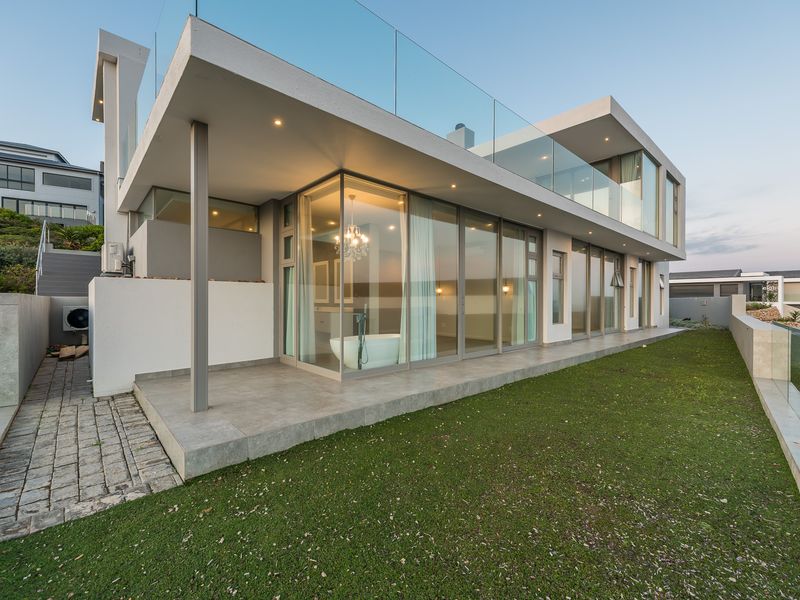 Modern architectural exterior rear view featuring a spacious patio, glass balustrades, and large floor-to-ceiling windows overlooking a landscaped garden area.