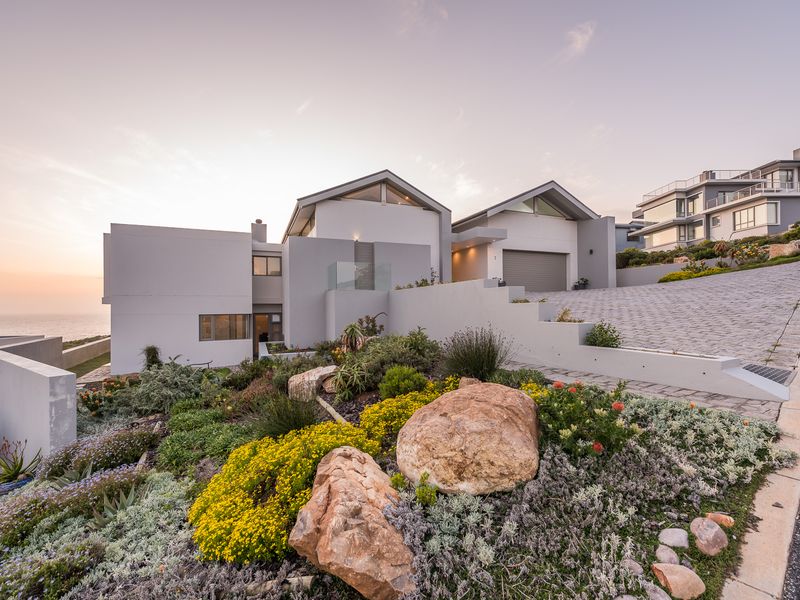 Modern architectural house exterior featuring a paved driveway, landscaped garden with rock features, and a double garage, set against a coastal backdrop.