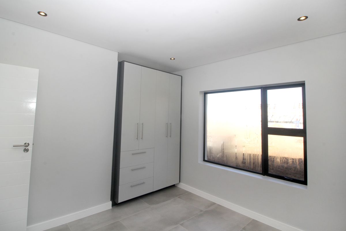 A clean, modern bedroom featuring neutral tiled flooring, built-in cupboards, and recessed downlighting, offering a bright and minimalist space.