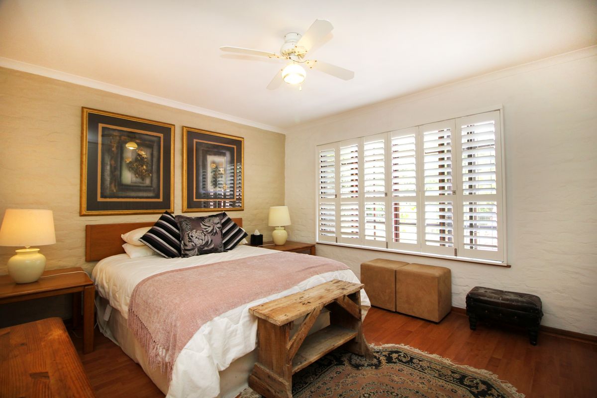 A well-maintained bedroom featuring warm wood flooring, textured walls, and white plantation shutters, complemented by a central ceiling fan.