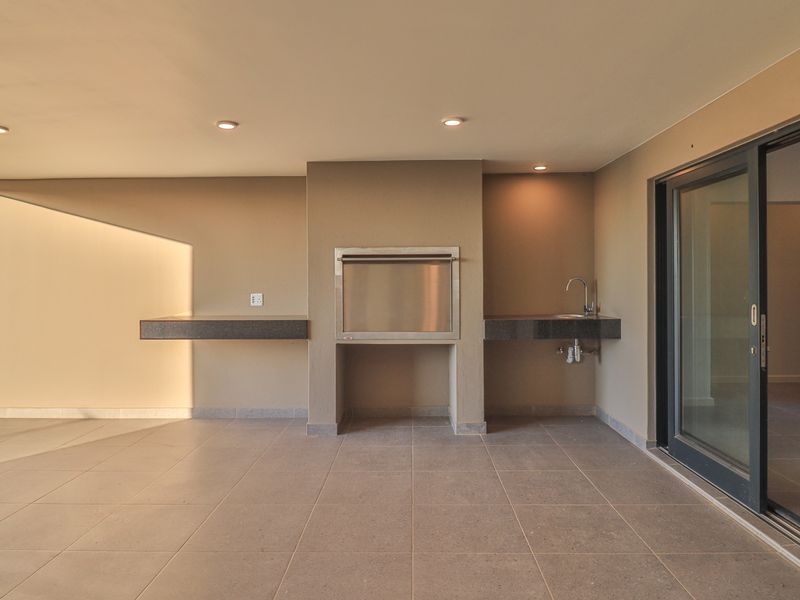 A modern covered patio featuring a built-in braai, stone prep counters with a sink, recessed lighting, and large sliding glass doors.