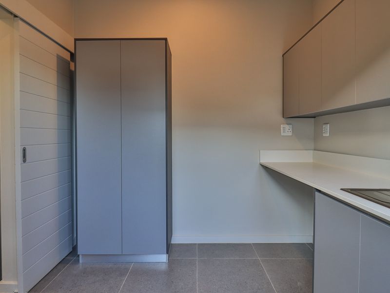 A modern, clean laundry room featuring sleek grey built-in cabinetry, a white countertop with an integrated sink, and neutral tiled flooring.