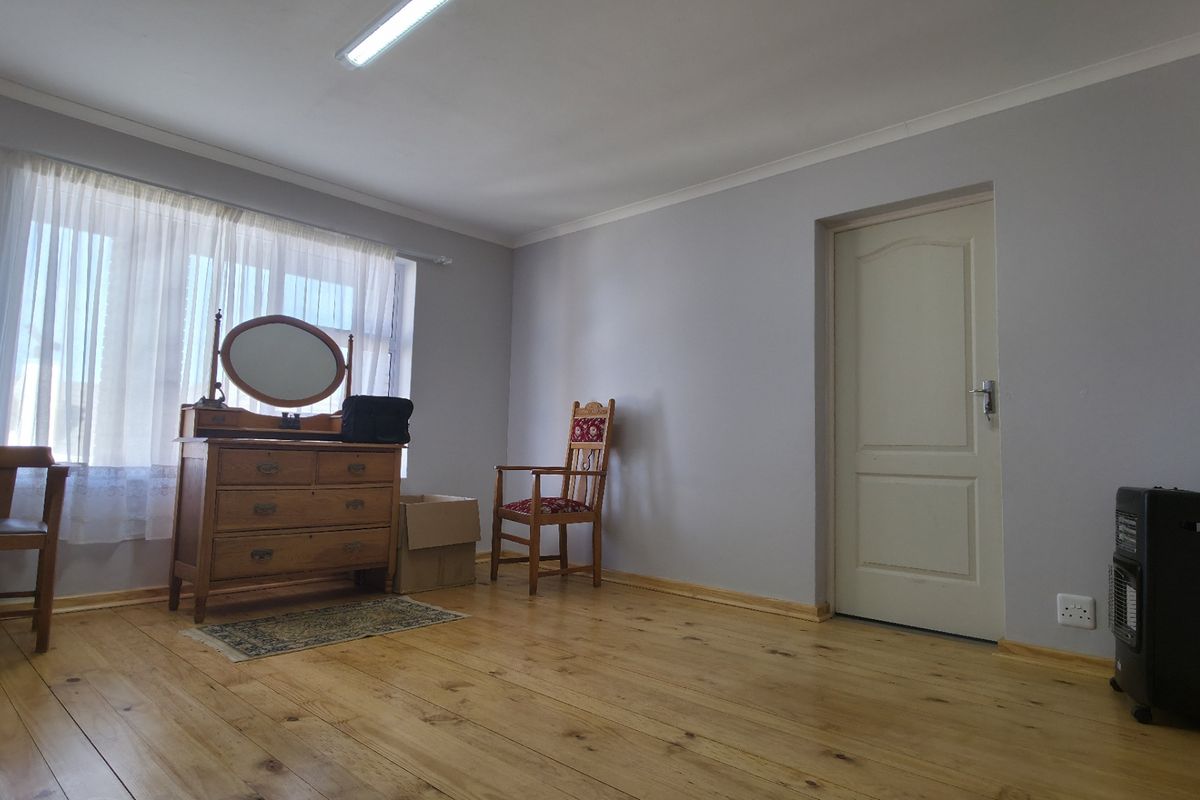 A bedroom featuring polished wooden flooring, neutral grey walls, and a large window allowing natural light, providing a clean and functional living space.