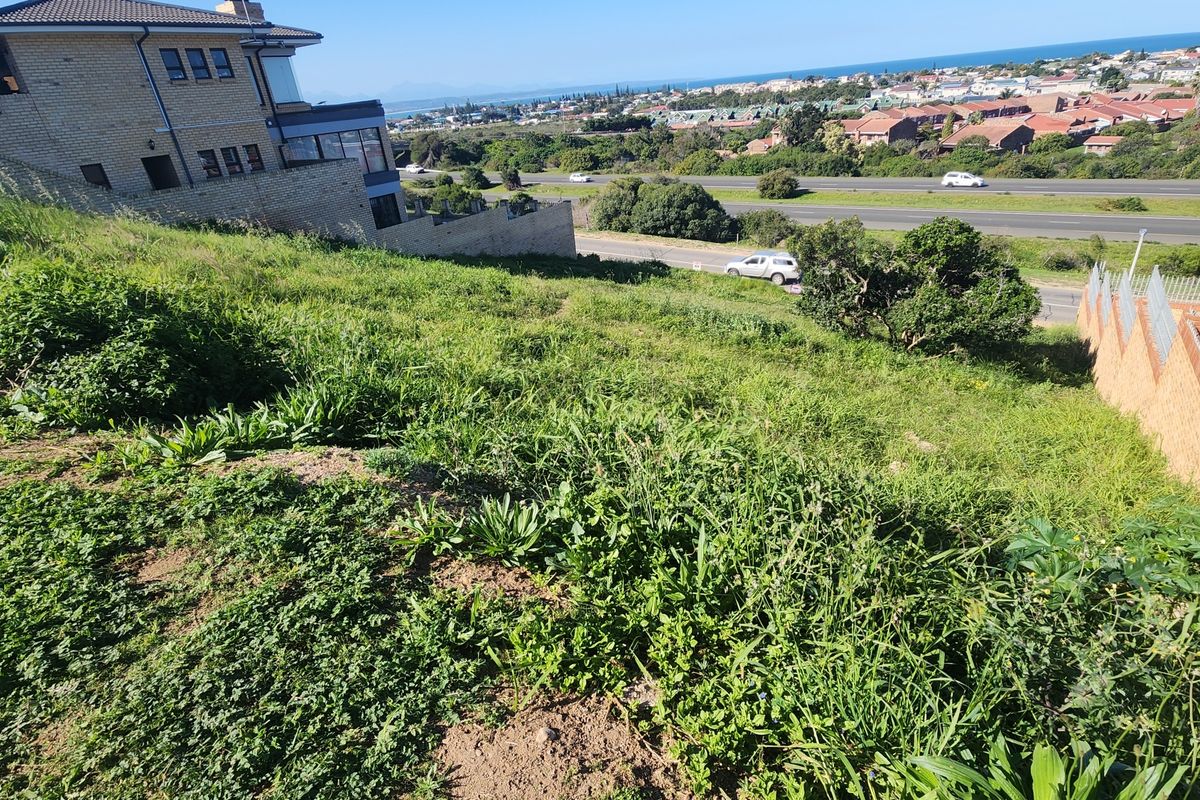An elevated view of a sloped residential vacant land plot featuring natural vegetation and adjacent retaining walls with distant coastal views.