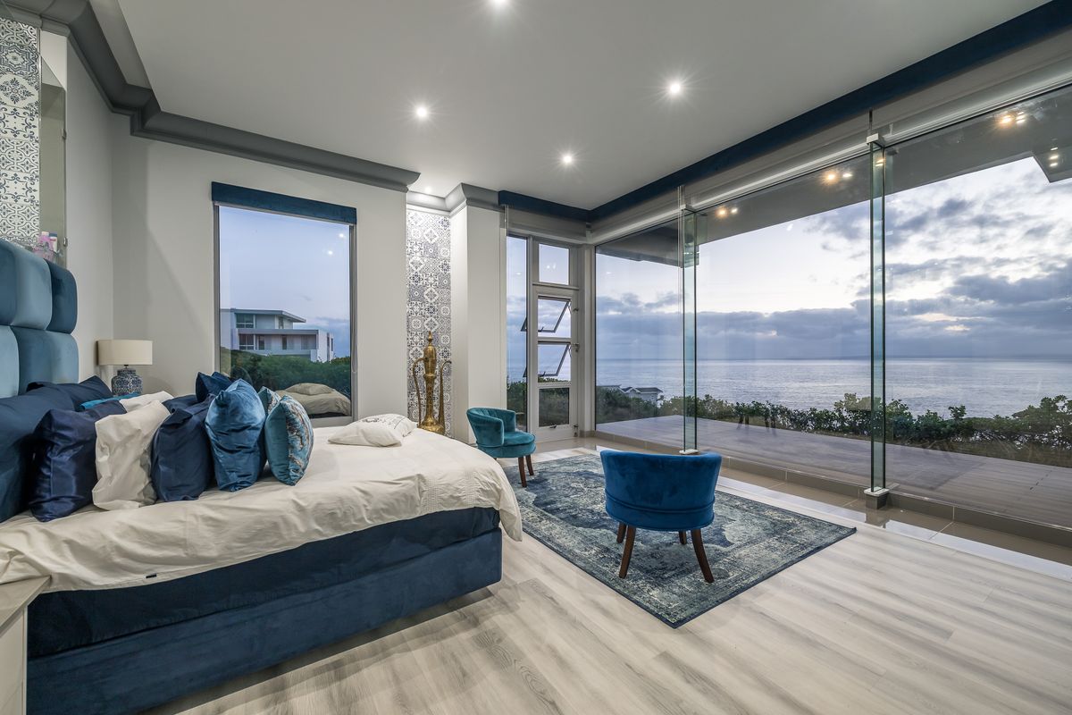 Spacious modern bedroom featuring floor-to-ceiling glass walls with panoramic ocean views, high-end finishes, and direct access to an outdoor deck.