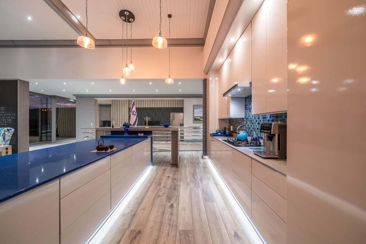 Modern kitchen featuring a large blue quartz island, high-gloss cabinetry, mosaic tile backsplash, and integrated under-cabinet lighting with a seamless open-plan flow.