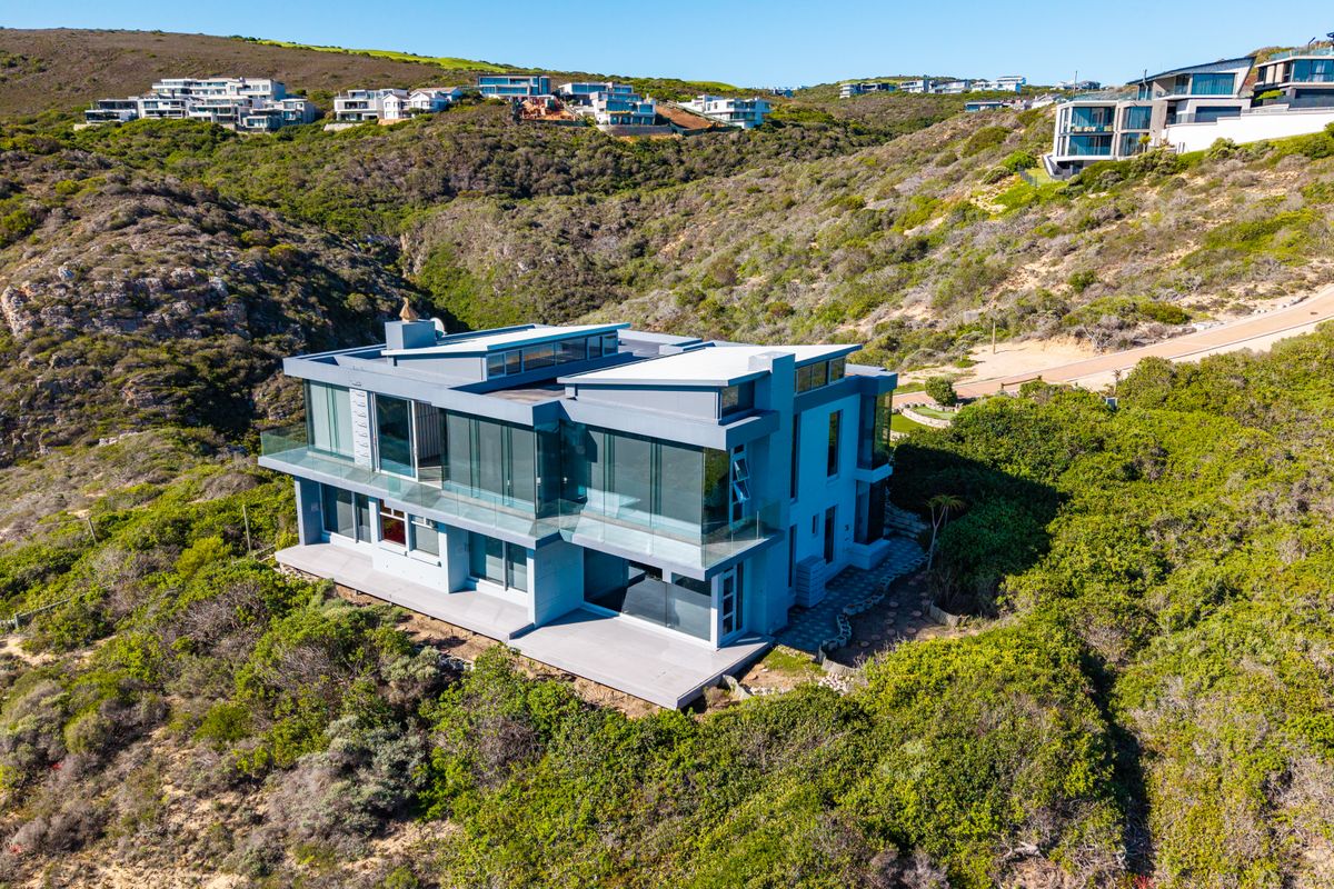Aerial view of a modern multi-level architectural home featuring extensive glass facades, flat roof design, and surrounding natural coastal landscape.