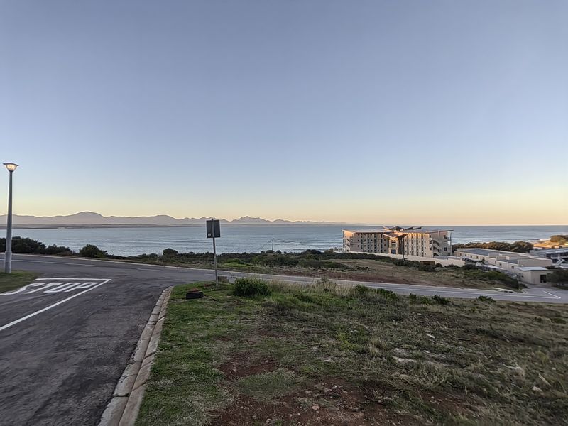 A scenic view overlooking the coastline and ocean, featuring a paved road in the foreground and distant mountain ranges under a clear sky.