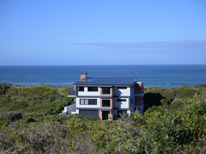 An elevated aerial view of a modern multi-story coastal home featuring brick and rendered finishes, a metal roof, and a double garage.