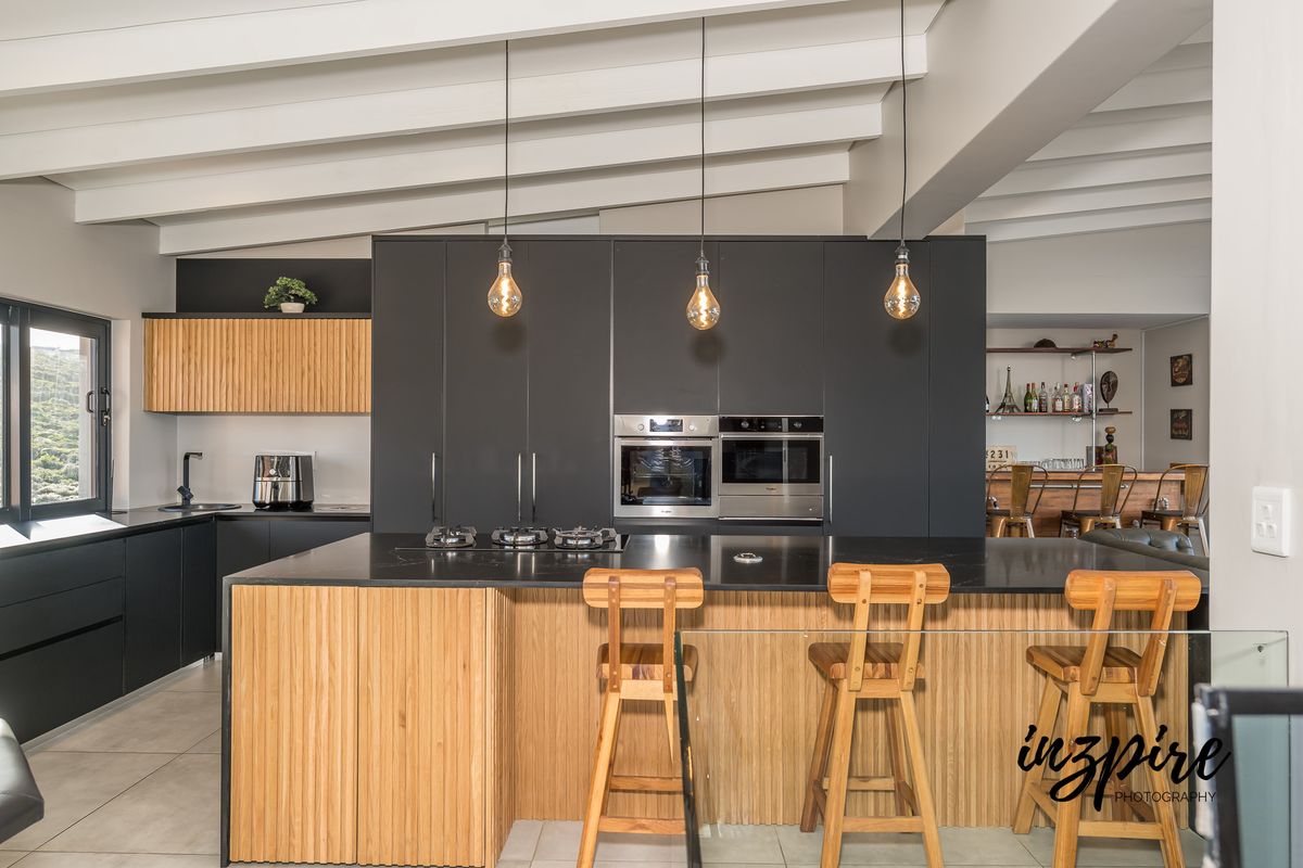 Modern kitchen featuring a large island with stone countertops, integrated appliances, sleek dark cabinetry, and exposed white ceiling beams.