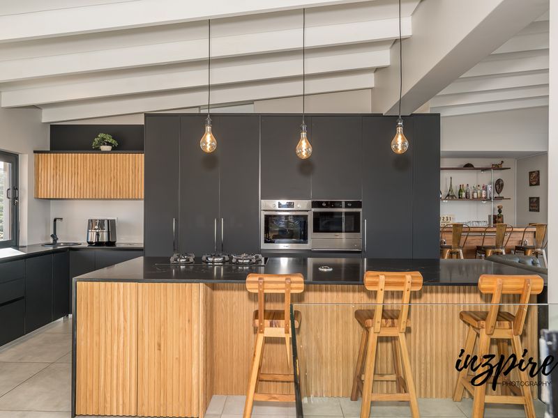 Modern kitchen featuring a large island with stone countertops, integrated appliances, sleek dark cabinetry, and exposed white ceiling beams.
