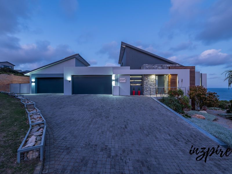 Modern architectural home featuring a double garage, stone and timber facade accents, and a paved driveway, set against a coastal backdrop at dusk.