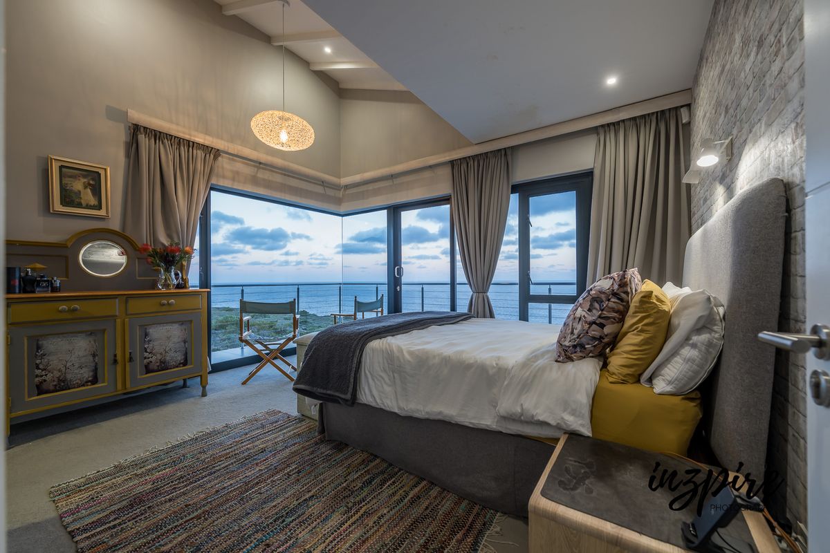 A modern bedroom featuring a vaulted ceiling, exposed brick wall, and expansive corner windows offering panoramic ocean views.