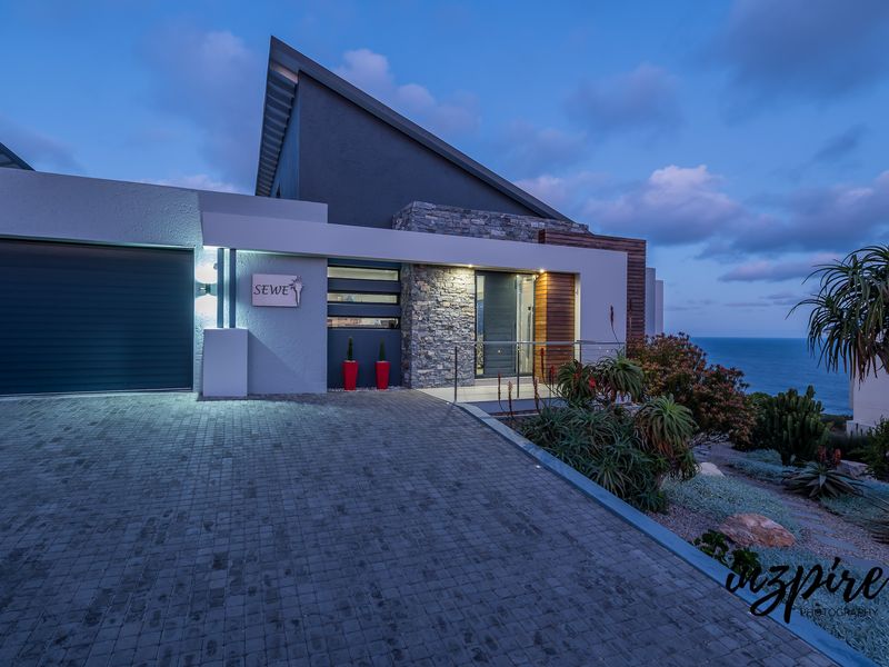Modern architectural front facade featuring stone cladding, a paved driveway, and a sleek garage entrance with ocean views at dusk.