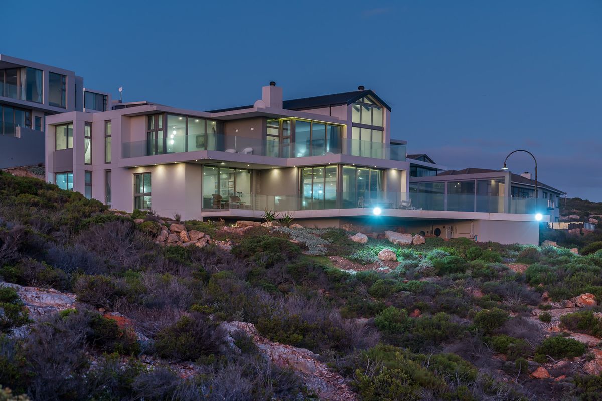 A modern multi-level architectural home featuring expansive glass facades, integrated balconies, and contemporary exterior lighting, set against a natural coastal landscape.