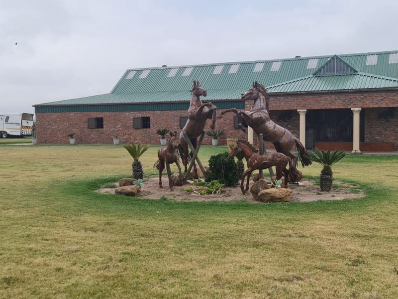 A spacious farm property featuring a brick building with a metal roof, a covered porch, and a well-maintained garden with a central horse sculpture.