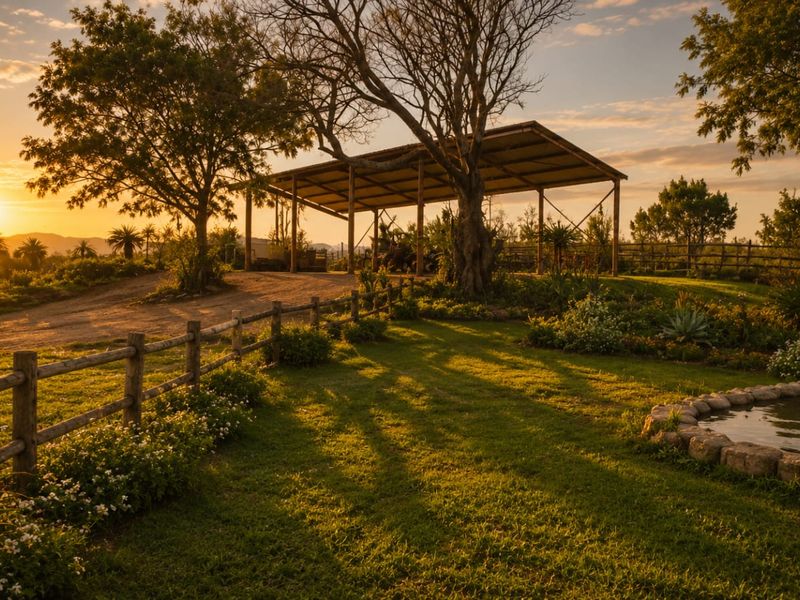 A scenic garden view featuring a stone-edged pond, timber fencing, and an open-sided barn structure set against a sunset landscape.