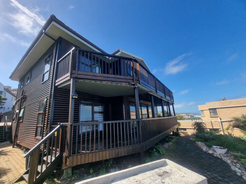 A two-story timber-clad house featuring a spacious wrap-around balcony with wooden balustrades and sliding glass doors, set in a landscaped garden area.