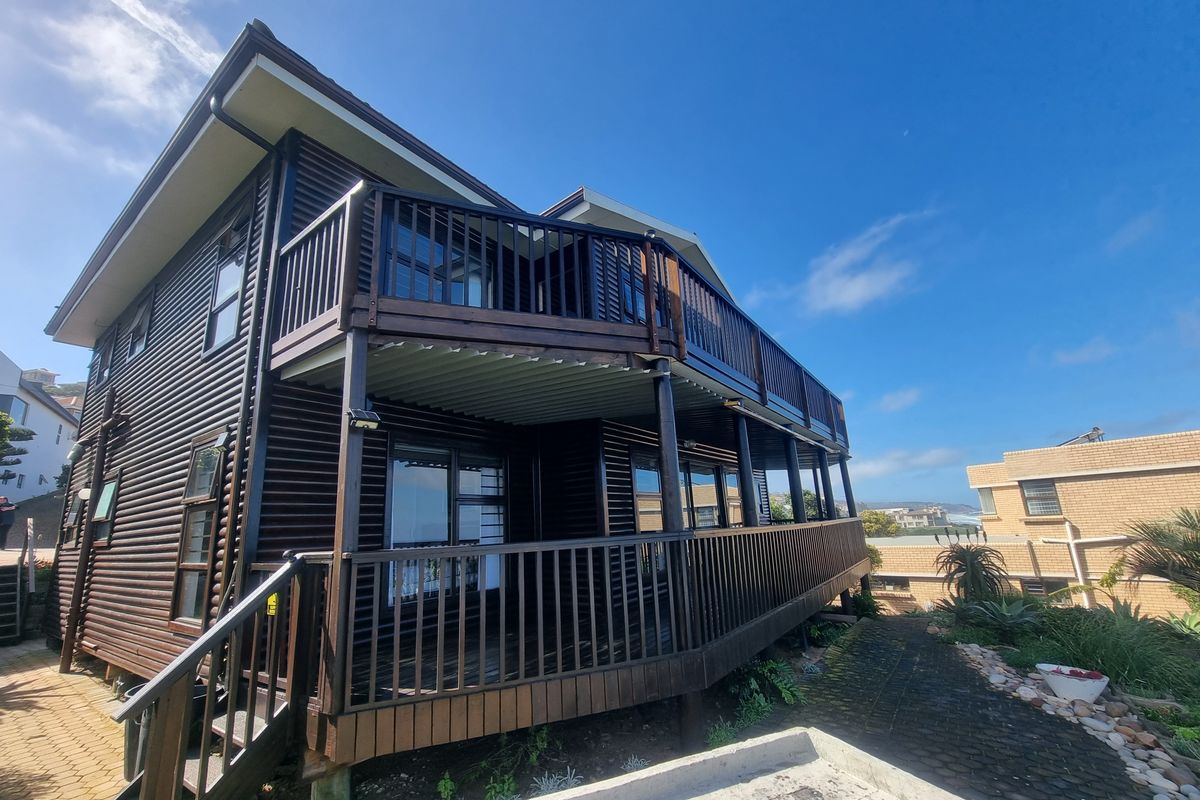 A two-story timber-clad house featuring a spacious wrap-around balcony with wooden balustrades and sliding glass doors, set in a landscaped garden area.