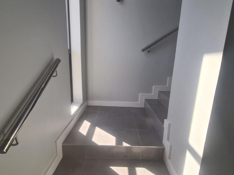 A clean, modern staircase featuring neutral grey floor tiles and a sleek stainless steel handrail, illuminated by natural light from a side window.