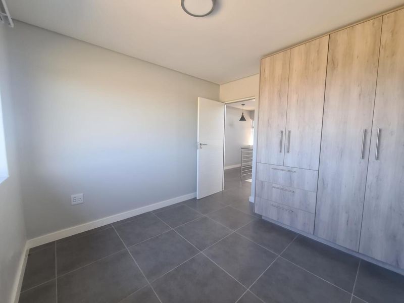 A clean, modern bedroom featuring neutral grey floor tiles, light wood-finish built-in cupboards, and a minimalist aesthetic.