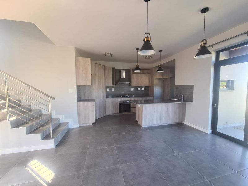 Modern open-plan kitchen featuring light wood cabinetry, integrated appliances, and a breakfast island, seamlessly connected to a tiled living area with a staircase.