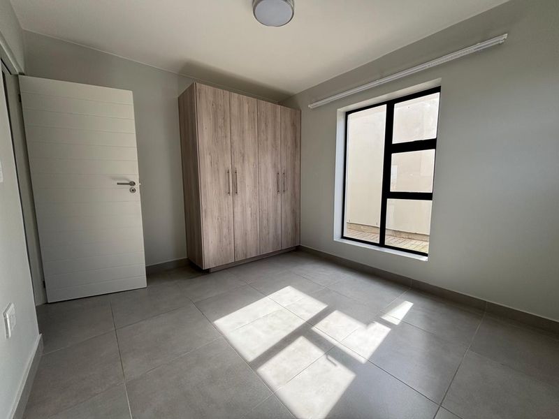A bright, modern bedroom featuring neutral tiled flooring, a built-in wooden wardrobe, and large aluminium-framed windows providing natural light.