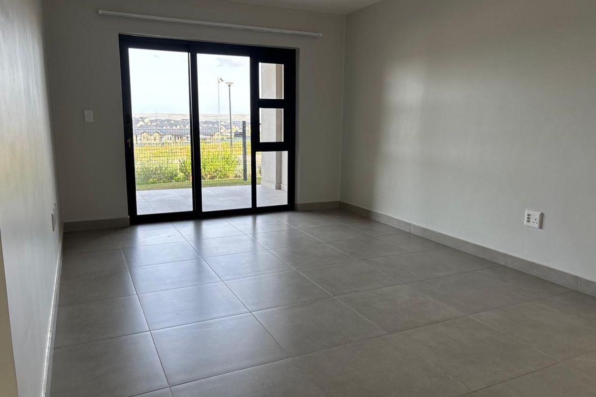 A bright, empty living room featuring modern grey floor tiles and a large sliding glass door providing access to an outdoor patio area.