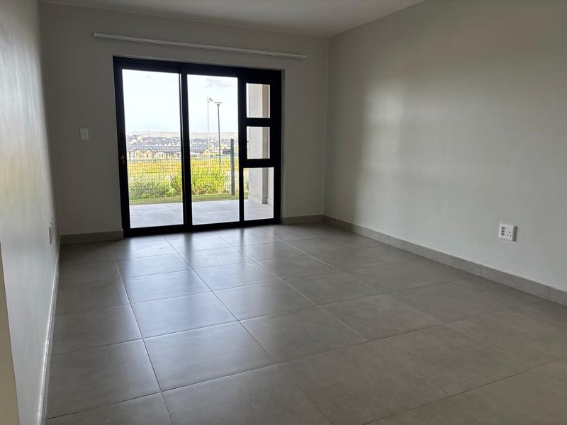 A bright, empty living room featuring modern grey floor tiles and a large sliding glass door providing access to an outdoor patio area.