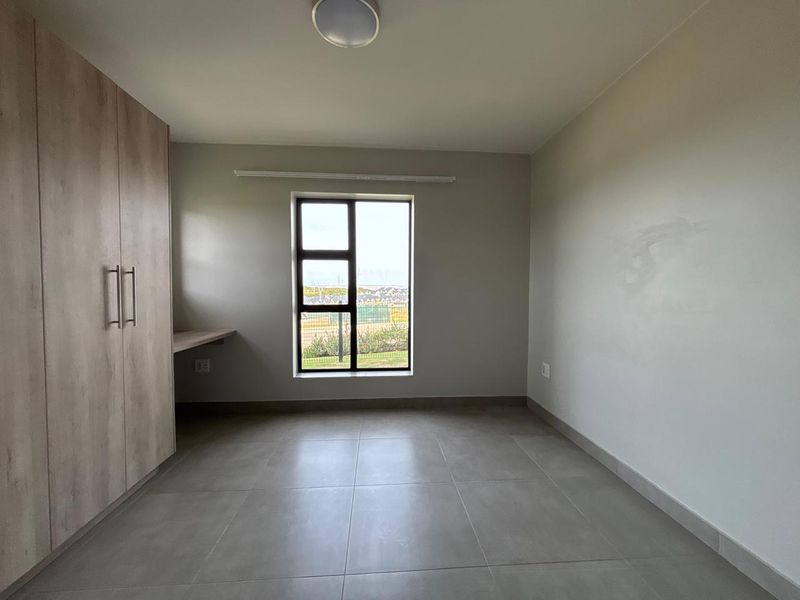 A clean, empty bedroom featuring neutral tiled flooring, built-in cupboards, a integrated desk, and a window providing natural light.