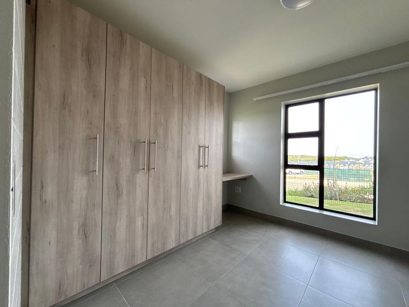 A bright, modern bedroom featuring light wood-finish built-in cupboards, a matching built-in desk, neutral tiled flooring, and a large window with exterior views.
