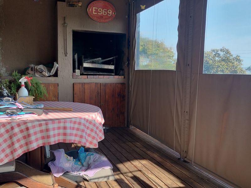Covered patio area featuring timber decking, an exposed brick wall, and a built-in braai, enclosed with retractable canvas blinds for all-weather use.