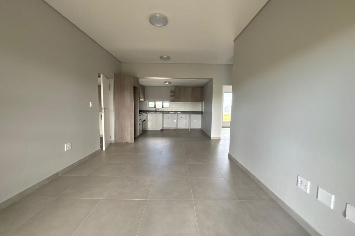 An open-plan living and kitchen area featuring neutral tiled flooring, modern cabinetry, and a clean subway tile backsplash in a bright, vacant space.
