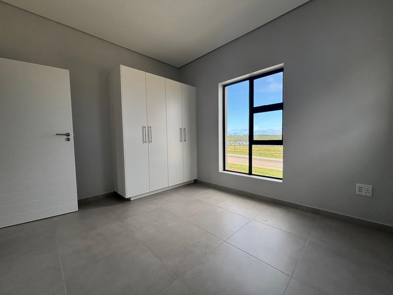 A bright, modern bedroom featuring neutral tiled flooring, built-in cupboards, and a large window with scenic exterior views.