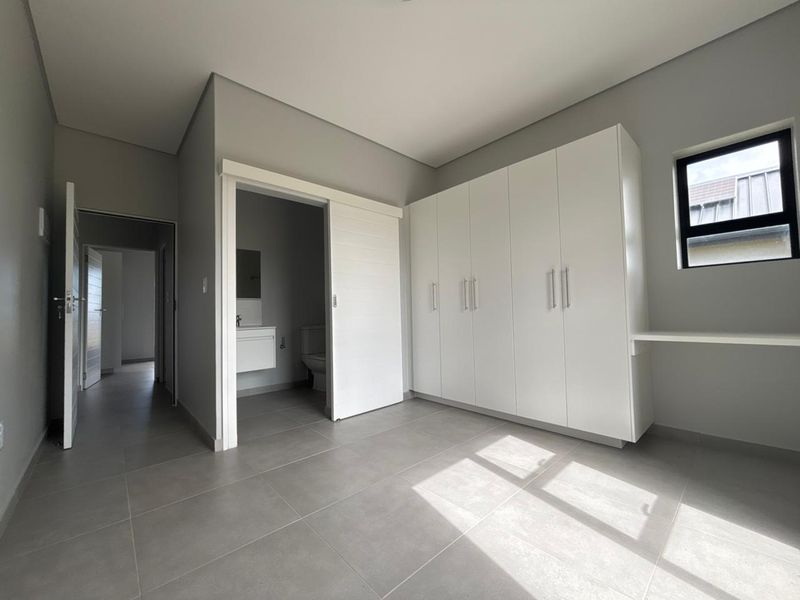 A modern, empty bedroom featuring neutral grey floor tiles, built-in white cupboards, a built-in desk, and an en-suite bathroom with a sliding door.