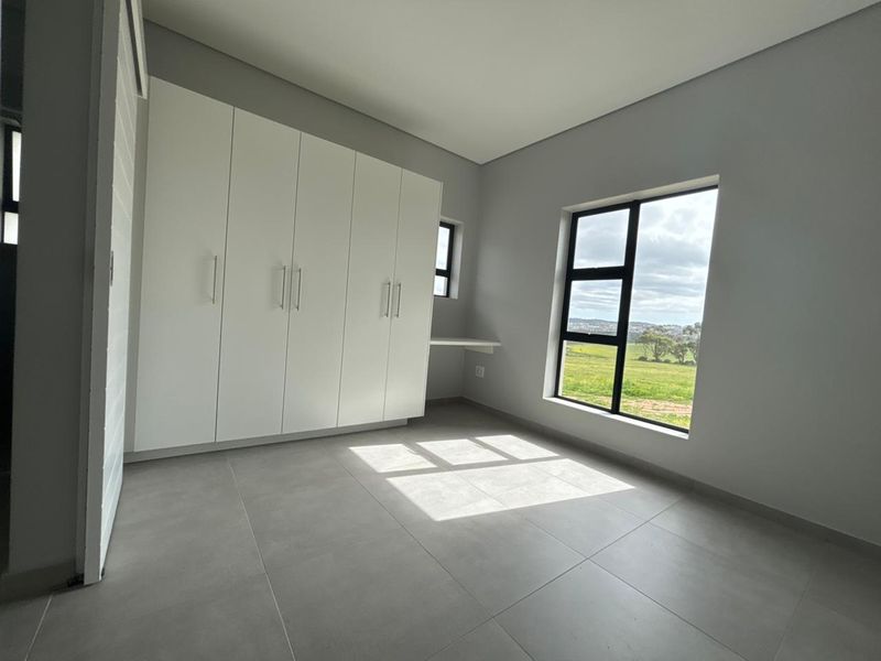 A bright, modern bedroom featuring neutral grey floor tiles, ample built-in cupboard space, a built-in desk, and large windows with scenic views.