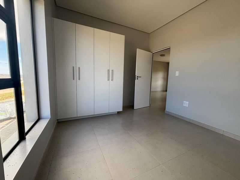 A bright, modern bedroom featuring neutral tiled flooring, a large window for natural light, and a spacious built-in wardrobe.