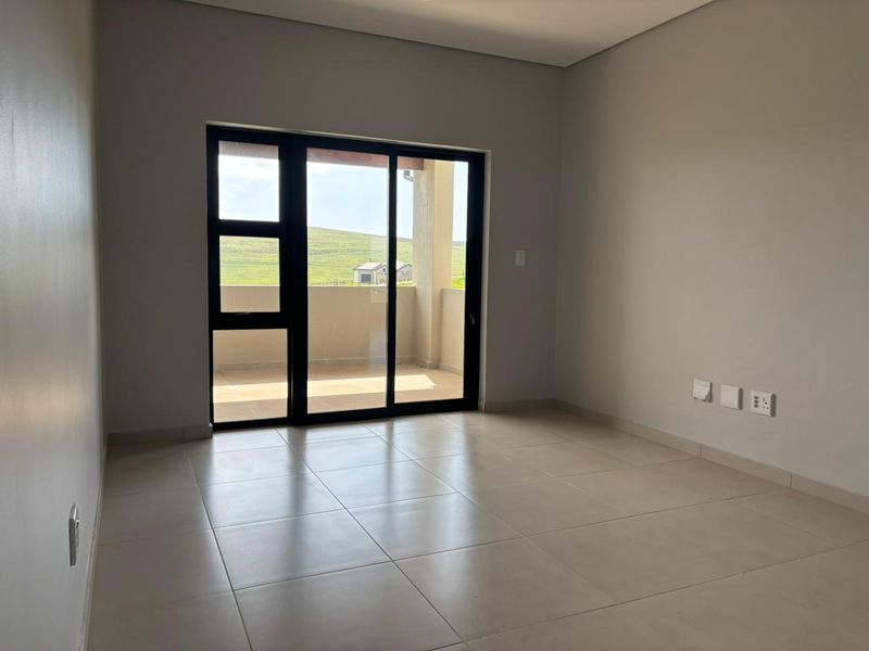A bright, empty living room featuring neutral tiled flooring and a large sliding glass door that opens onto a private balcony.
