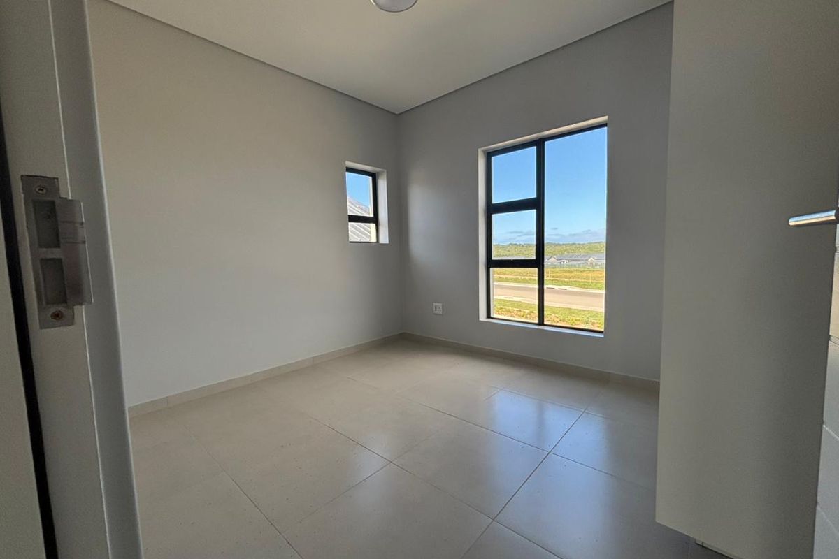 A bright, empty bedroom featuring neutral tiled flooring, modern aluminium window frames, and clean, minimalist finishes with a view of the surrounding area.