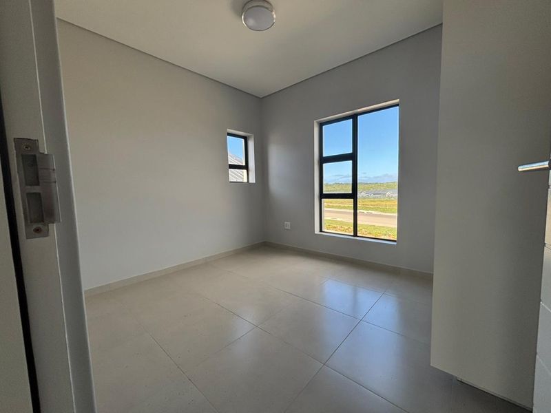 A bright, empty bedroom featuring neutral tiled flooring, modern aluminium window frames, and clean, minimalist finishes with a view of the surrounding area.