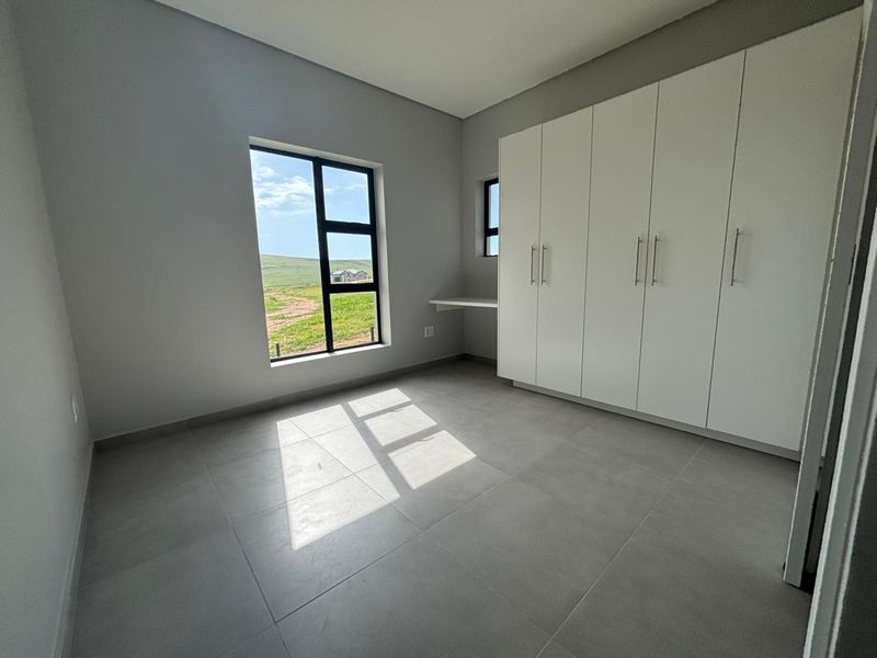 A bright, modern bedroom featuring neutral tiled flooring, built-in white cupboards, a built-in desk, and large windows with views of the surrounding landscape.