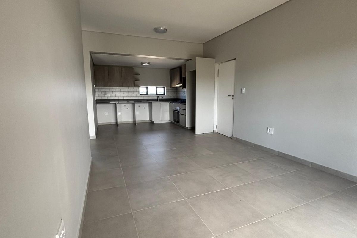 An open-plan living and kitchen area featuring neutral tiled flooring, modern cabinetry, subway tile backsplash, and integrated kitchen appliances in a clean, vacant space.