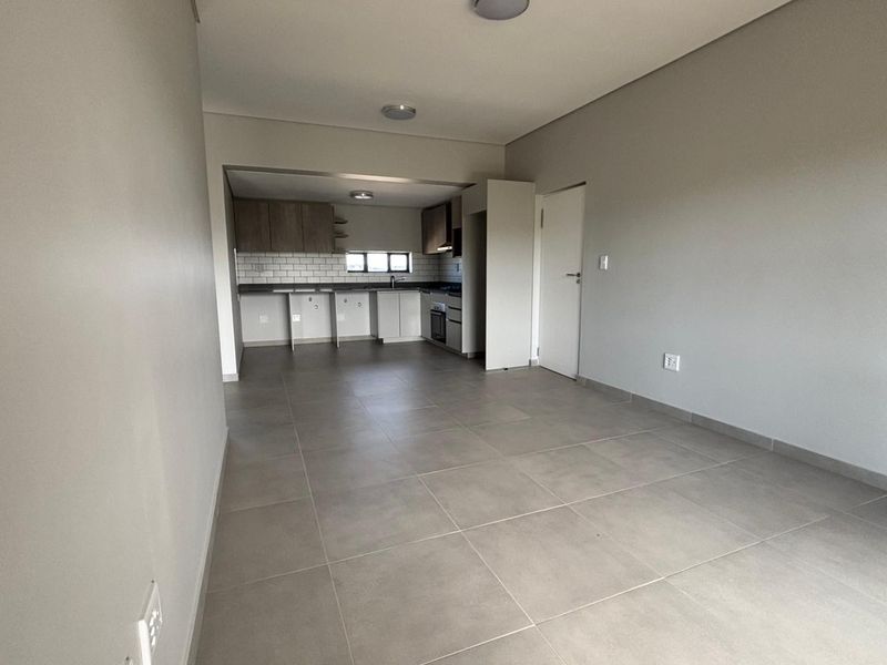 An open-plan living and kitchen area featuring neutral tiled flooring, modern cabinetry, subway tile backsplash, and integrated kitchen appliances in a clean, vacant space.