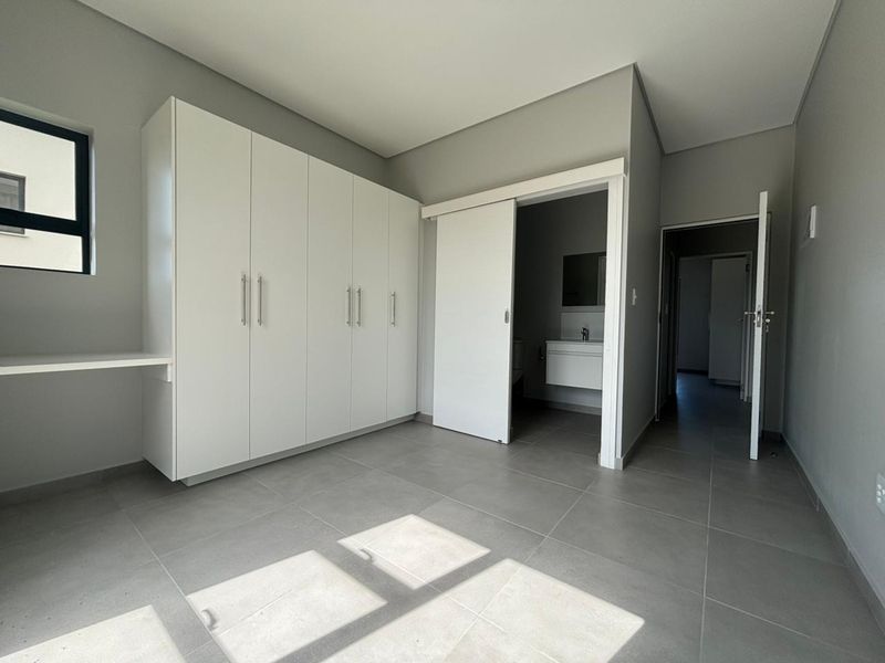 A modern, empty bedroom featuring neutral tiled flooring, built-in white cupboards with an integrated desk, and direct access to an en-suite bathroom.