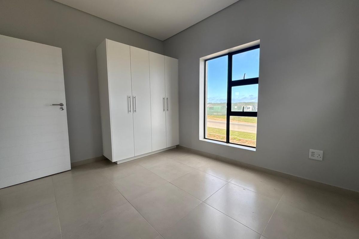 A bright, modern, and empty bedroom featuring neutral tiled flooring, built-in white cupboards, and a large window with natural light.