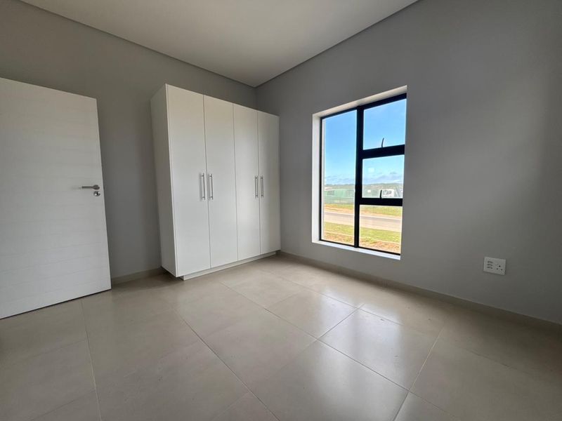 A bright, modern, and empty bedroom featuring neutral tiled flooring, built-in white cupboards, and a large window with natural light.