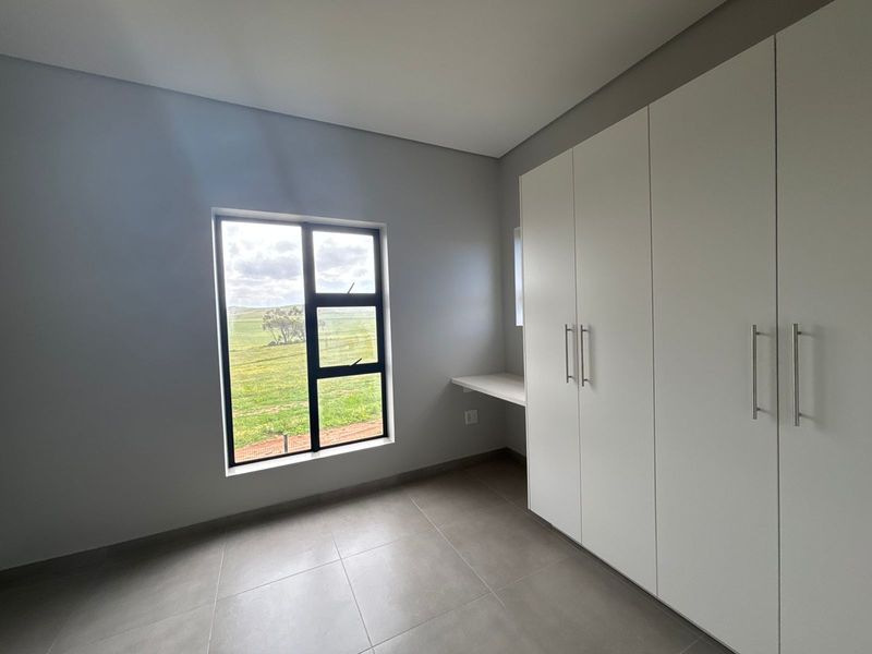 A bright, modern bedroom featuring neutral tiled floors, ample built-in cupboard space, a built-in desk, and large windows with scenic views.