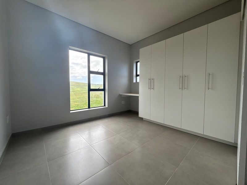 A bright, modern bedroom featuring neutral tiled flooring, ample built-in cupboard space, and a convenient integrated desk area with scenic views.