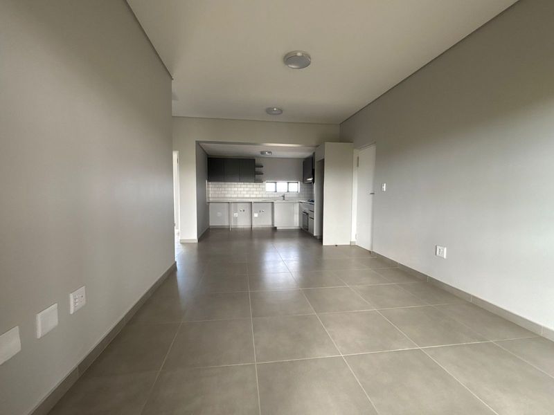 An open-plan living and kitchen area featuring neutral tiled flooring, modern cabinetry, and a subway tile backsplash in a clean, vacant space.